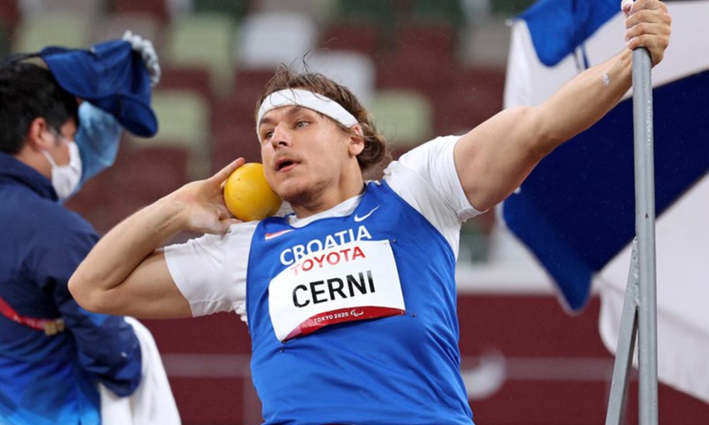 Na slici je para atletičar koji baca kuglu jednom rukom dok se drugom drži za metalnu šipku. Atletičar nosi bijelu majicu s plavim markerom na kojem piše 'Cerni'.