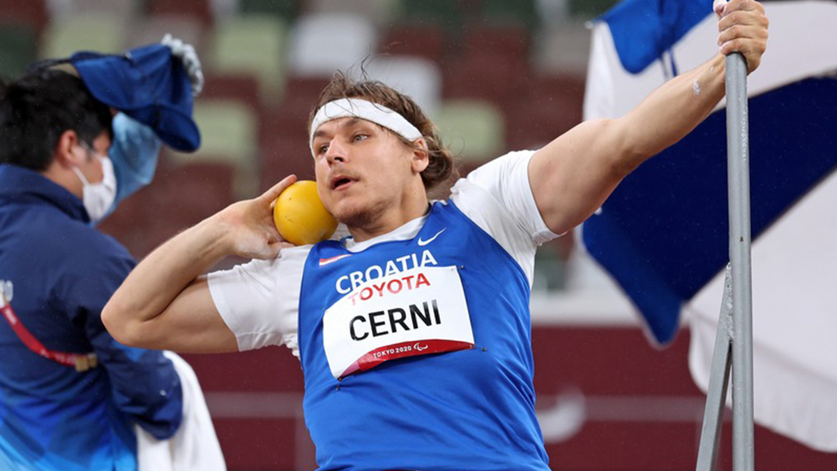 Na slici je para atletičar koji baca kuglu jednom rukom dok se drugom drži za metalnu šipku. Atletičar nosi bijelu majicu s plavim markerom na kojem piše 'Cerni'.