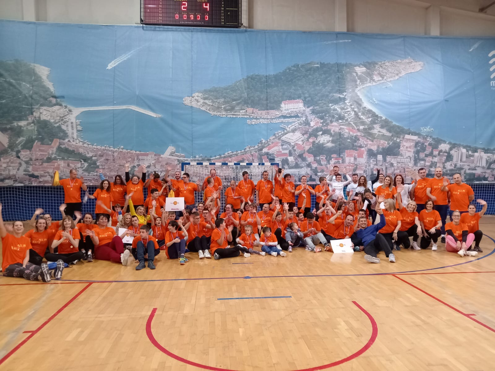 Na slici je velika grupa ljudi u narančastim majicama u sportskoj dvorani. Grupa pozira za fotografiju pred plavom pozadinom s otisnutim gradskim pejzažem. Pozadina je obješena na koš za košarku. Na njoj je prikazan grad s neboderima, mostovima i rijekom.