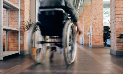 Ovo je slika osobe u invalidskim kolicima koja se kreće hodnikom. Hodnik ima drveni pod i zidove od opeke. U pozadini su biljke i namještaj. Slika je snimljena iz niskog kuta i osoba je u pokretu, stvarajući zamagljen učinak.