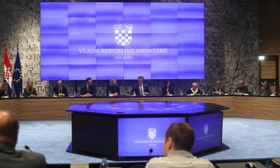 Ovo je slika konferencijske dvorane Vlade Republike Hrvatske. Na velikom plavom ekranu je grb Hrvatske i natpis “Vlada Republike Hrvatske”. U dvorani je veliki okrugli stol s mikrofonima i natpisima. Za stolom sjede ljudi.