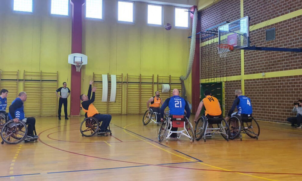 Ovo je fotografija košarkaške utakmice u dvorani. Na slici se vide igrači u invalidskim kolicima koji igraju košarku. Dvorana ima drveni pod i žute zidove. Na lijevoj strani slike nalazi se košarkaški obruč. Na desnoj strani slike nalaze se gledatelji. Slika je snimljena s perspektive bočne linije.