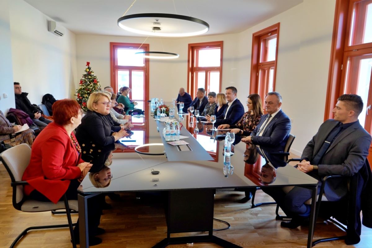 Na slici je prikazana soba u kojoj se održava sastanak. Osobe su smještene oko dugog stola.U pozadini se vidi božićno drvce, što ukazuje na to da je slika snimljena tijekom blagdanske sezone.