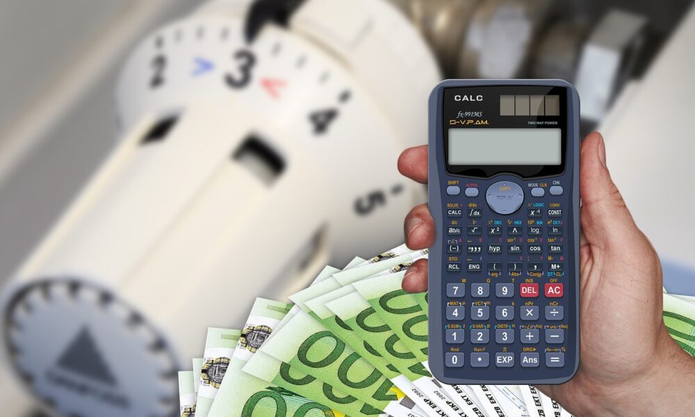 Na slici se nalazi termostatski ventil, novčanice po sto eura i vidi se ruka u kojoj je kalkulator. Sve zajedno ilustrira skupoću i energetsku krizu.