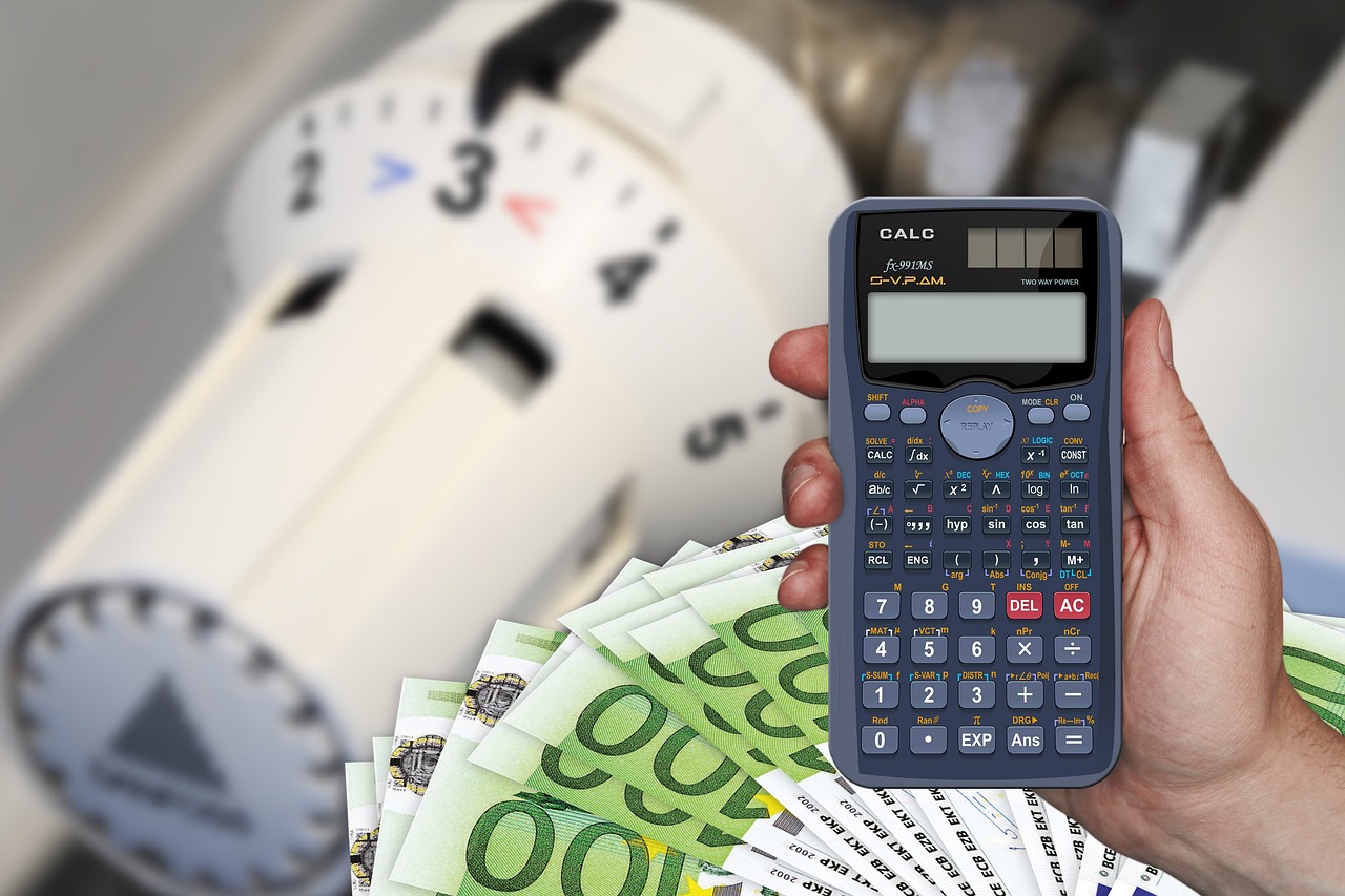 Na slici se nalazi termostatski ventil, novčanice po sto eura i vidi se ruka u kojoj je kalkulator. Sve zajedno ilustrira skupoću i energetsku krizu.