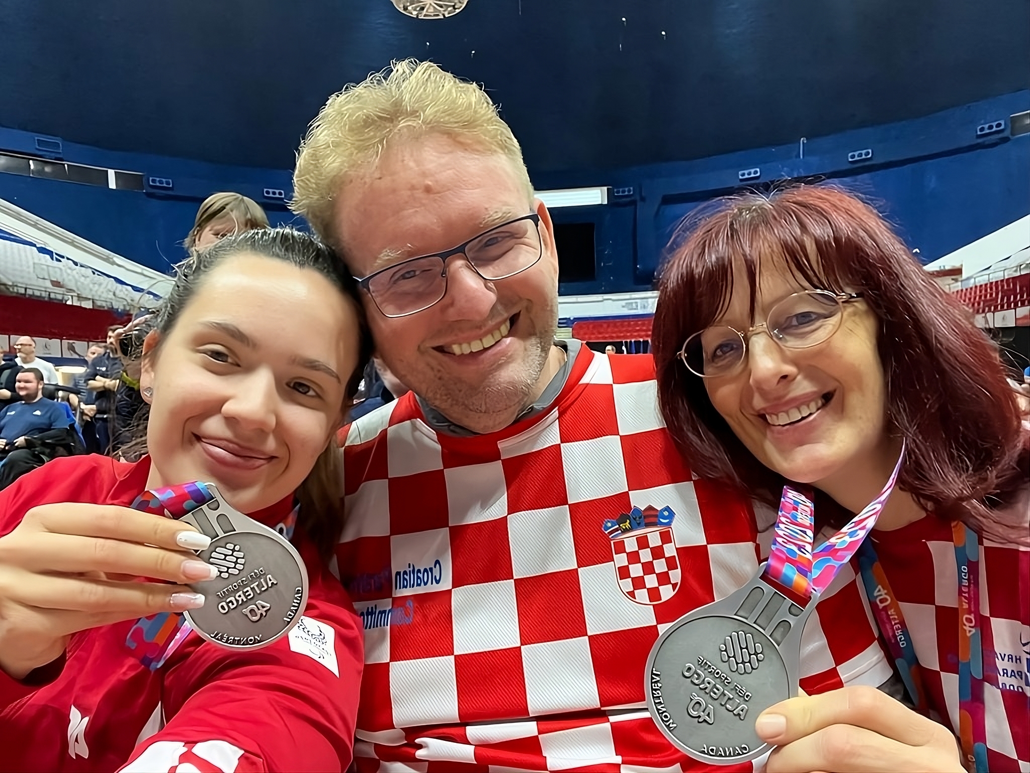 Na ovoj slici su tri osobe, a dvije drže medalje. Njihova odjeća ima šahovnicu crvene i bijele boje. Medalje su velike i okrugle, s natpisom koji nije jasno vidljiv. Pozadina prikazuje unutrašnjost sportske arene s plavim sjedalicama.