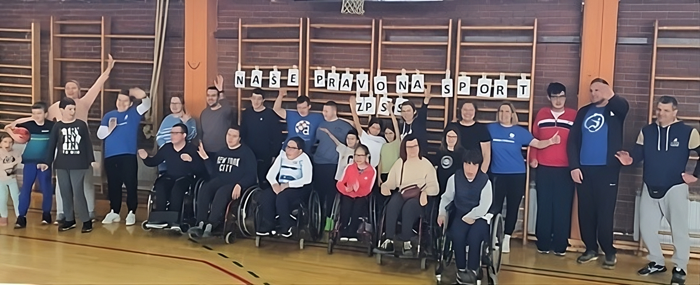 Na slici je grupa ljudi u sportskoj dvorani. Neki od njih su u kolicima, dok drugi stoje. Iza njih je transparent s tekstom.