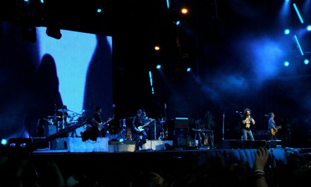 Ova slika je fotografija koncertne pozornice s bendom koji nastupa. Pozornica je osvijetljena plavim i bijelim svjetlima koja stvaraju kontrast s tamnom pozadinom. Pozadina pozornice je veliki ekran. Publika je vidljiva u prvom planu.
