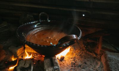 Ovo je fotografija velikog crnog lonca s ručkom iznad otvorenog plamena. Lonac je pun variva koje izgleda da vrije. Vatra je napravljena od malih kamenja i grančica. Pozadina je tamni, prljavi zid s drvenim gredama. Iz lonca i vatre izlazi dim.