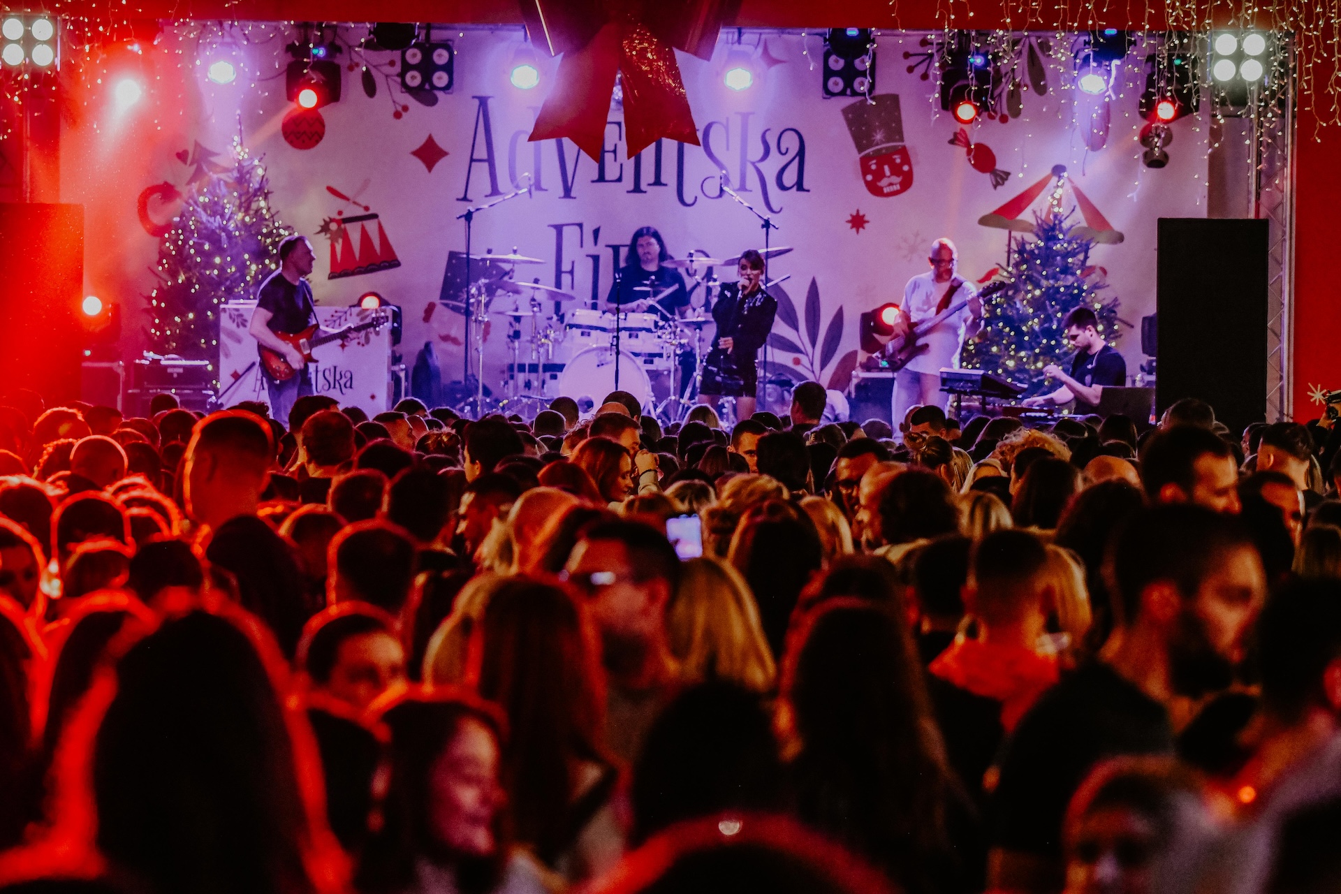 Ova slika prikazuje koncertnu atmosferu tijekom božićnog razdoblja. Na pozornici je bend koji svira, okružen božićnim dekoracijama i osvjetljenjem. Publika je u prvom planu. Pozadina pozornice sadrži veliki tekst i razne simbole vezane uz blagdane. Rasvjeta je živahna i doprinosi svečanom ozračju događaja.