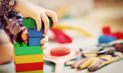Na fotografiji su dječje ruke koje slažu šarene plastične kocke. U pozadini se vide različite igračke i predmeti rasuti po podu.