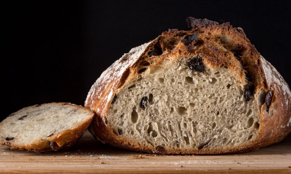 Na slici je veliki komad kruha koji je djelomično narezan. Kriška kruha leži pored glavnog dijela na drvenoj površini, a pozadina je tamna.