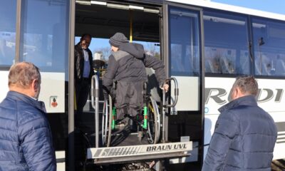 Ovo je fotografija osobe u invalidskim kolicima koja koristi dizalo za invalidska kolica kako bi se ukrcala u autobus. Na fotografiji se nalaze još dvije osobe, jedna pomaže osobi u invalidskim kolicima, a druga stoji u blizini.