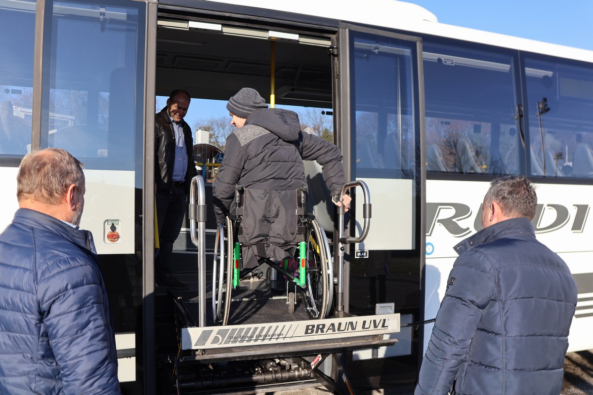Ovo je fotografija osobe u invalidskim kolicima koja koristi dizalo za invalidska kolica kako bi se ukrcala u autobus. Na fotografiji se nalaze još dvije osobe, jedna pomaže osobi u invalidskim kolicima, a druga stoji u blizini.