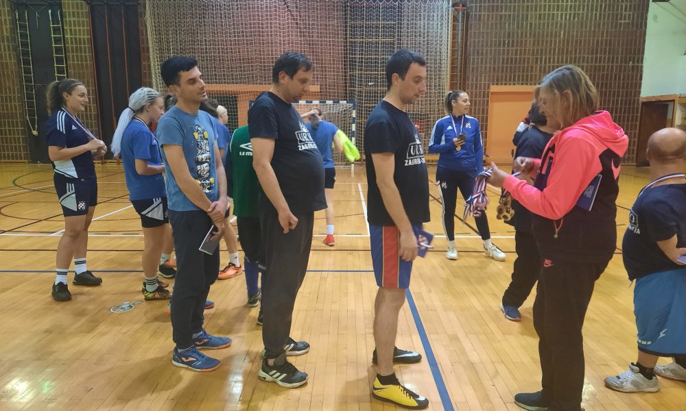 Na slici je grupa ljudi koji se bave futsalom u zatvorenom prostoru. Igrači nose različite boje dresova, hlačica i čarapa. Svi igrači nose sportske cipele. Pozadina je drveni pod (dvorana) s mrežom u pozadini.