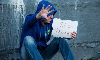 Ovo je slika muškarca koji sjedi na tlu ispred betonskog zida. Na sebi nosi plavu kapuljaču i traperice. Drži znak na kojem piše 'Stop drogama'. Pozadina se sastoji od betonskog zida s grafitima.