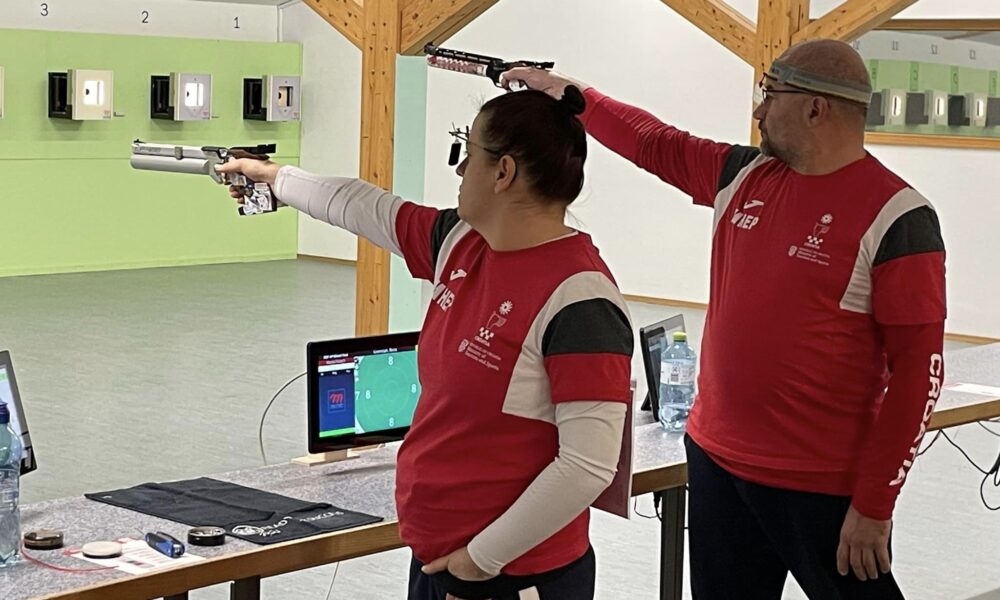 Ova slika prikazuje dvoje ljudi u crvenim majicama koji drže pištolje i ciljaju na mete u prostoriji sa zelenim zidovima i drvenim policama. Ljudi stoje jedan do drugog. Na stolu u pozadini nalazi se monitor računala i druga oprema.