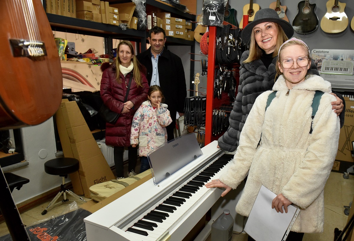 Fotografija prikazuje petero ljudi u trgovini punoj glazbenih instrumenata i opreme. Osobe su okupljene oko klavira.