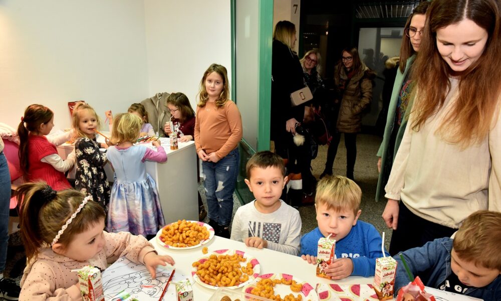 Na fotografiji je grupa djece i odraslih okupljenih oko stola s grickalicama. Djeca su zauzeta jedenjem i crtanjem, dok odrasli stoje u pozadini.