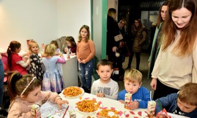 Na fotografiji je grupa djece i odraslih okupljenih oko stola s grickalicama. Djeca su zauzeta jedenjem i crtanjem, dok odrasli stoje u pozadini.