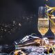 Ova slika prikazuje dvije čaše šampanjca, okružene zlatnim trakama i bočicom šampanjca na tamnoj pozadini sa svjetlucavim efektom. Zlatne vrpce se vijore oko čaša. Pozadina je tamna, ali osvijetljena sjajnim svjetlima koja stvaraju bokeh efekt.