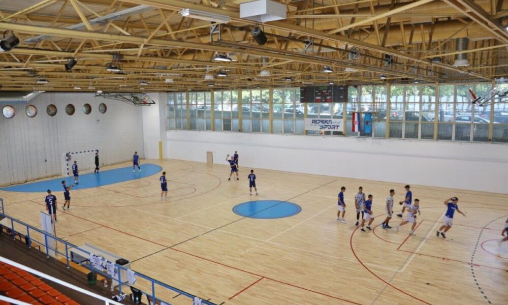 Ovo je slika sportske dvorane. Na slici se vidi drveni pod s oznakama za različite sportove i plavi centar. Igrači u plavim dresovima igraju neku vrstu sportske igre. Dvorana ima drvene stropne grede i velike prozore koji omogućuju prirodno svjetlo.