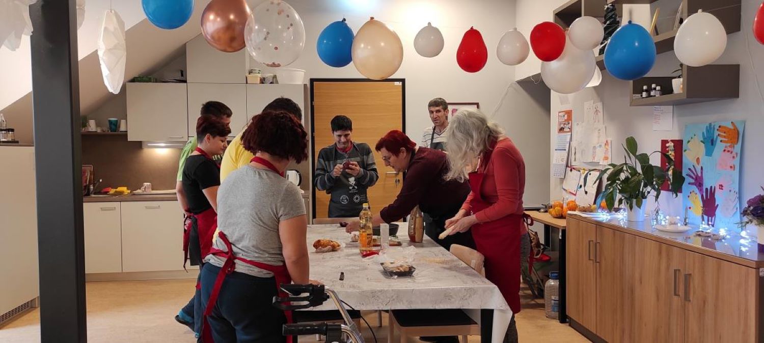 Na ovoj fotografiji se nalazi unutrašnji prostor, vjerojatno kuhinja, gdje se nekoliko ljudi okupilo. Prostor je ukrašen balonima različitih boja koji su pričvršćeni za strop. Ljudi se bave različitim aktivnostima oko stola i kuhinjskog pulta.