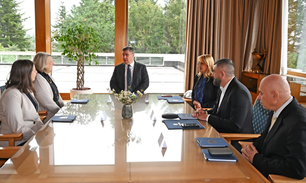 Ova slika prikazuje skupinu ljudi koja sjedi oko stola na sastanku. Oni su odjeveni u formalnu odjeću, uključujući odijela i poslovne haljine.
