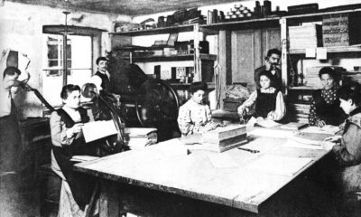 Ova slika prikazuje stari tiskarski ured s nekoliko osoba koje rade. U prostoriji je stari tiskarski stroj, a osobe su zauzete raznim aktivnostima povezanima s tiskanjem i pripremom materijala. Slika je crno-bijela i ima povijesnu vrijednost.