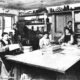 Ova slika prikazuje stari tiskarski ured s nekoliko osoba koje rade. U prostoriji je stari tiskarski stroj, a osobe su zauzete raznim aktivnostima povezanima s tiskanjem i pripremom materijala. Slika je crno-bijela i ima povijesnu vrijednost.