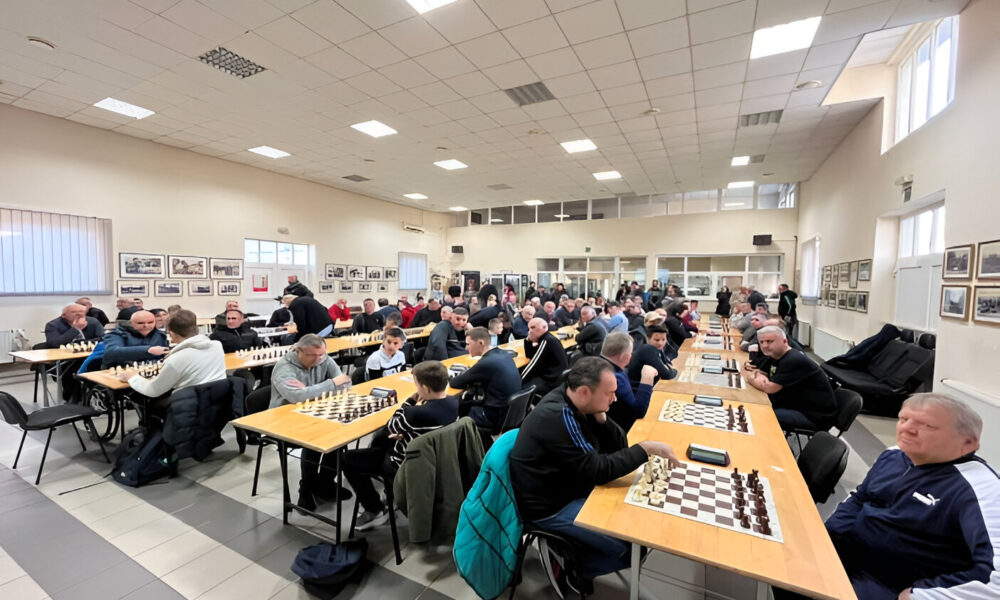 Ova slika prikazuje prostoriju u kojoj se održava šahovski turnir. Mnogi igrači sjede za dugim stolovima, a svaki stol ima šahovsku ploču. Prostorija je prostrana i dobro osvijetljena, s nekoliko prozora na zidu.