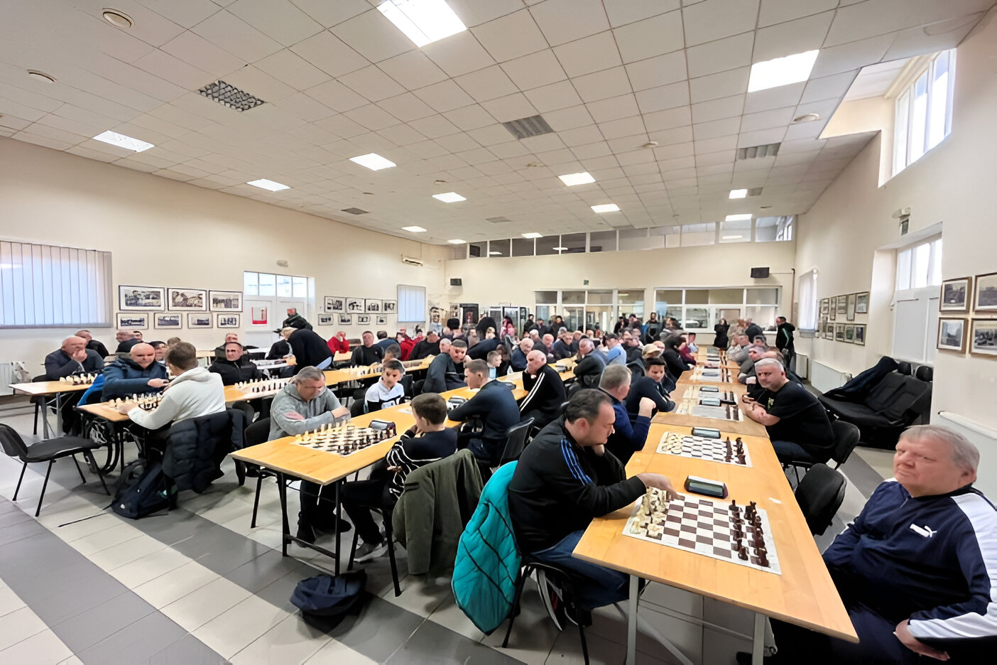 Ova slika prikazuje prostoriju u kojoj se održava šahovski turnir. Mnogi igrači sjede za dugim stolovima, a svaki stol ima šahovsku ploču. Prostorija je prostrana i dobro osvijetljena, s nekoliko prozora na zidu.