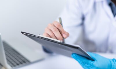 Na ovoj fotografiji vidimo osobu u bijelom laboratorijskom mantilu koja nosi plave medicinske rukavice i piše na kliničkom obrascu ili dokumentu. U pozadini se vidi laptop. Ova slika sugerira da se nalazimo u kliničkom okruženju, a osoba na slici vjerojatno obavlja neki medicinski posao.