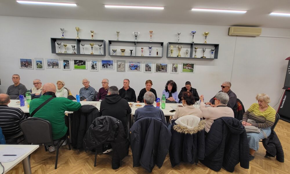 Ova slika prikazuje grupu ljudi koji sjede za stolom u prostoriji s bijelim zidovima. Na zidu iza njih nalazi se niz trofeja i fotografija. Prostorija izgleda kao neka vrsta klupske sobe ili sale za sastanke.