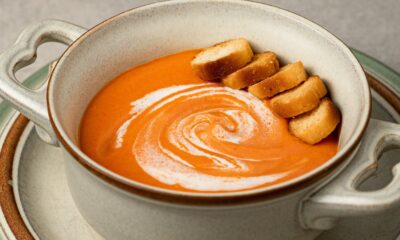 Na slici je zdjela kremaste juhe od tikve s nekoliko komadića prženog kruha i vrhnja na vrhu. Juha je narančaste boje i poslužena je u keramičkoj zdjeli s dvije ručke, koja stoji na tanjuru.