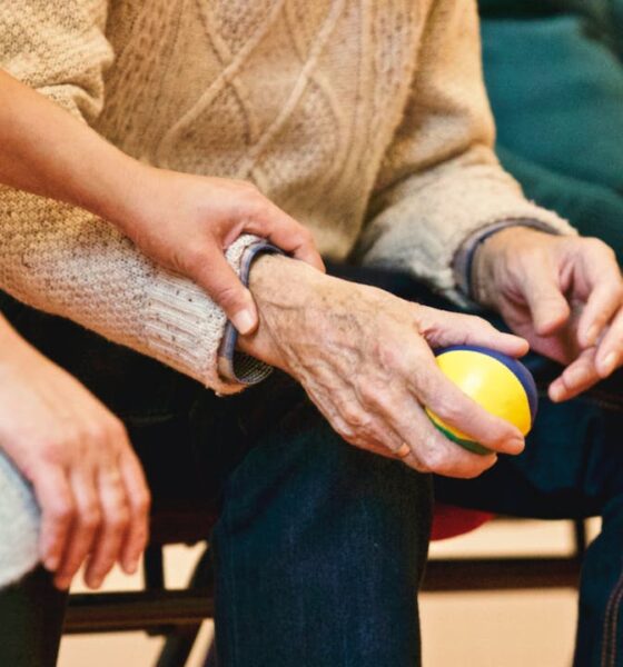 Slika prikazuje trenutak gdje osoba drži ruke starije osobe. Starija osoba drži malu lopticu sa žutim i plavim segmentima, možda za vježbu ili terapiju. Oboje su sjedeći, iako se u kadru vide samo njihova tijela i ruke. Nose ležernu odjeću; mlađa osoba u sivoj majici, a starija u bež džemperu i tamnim hlačama.
