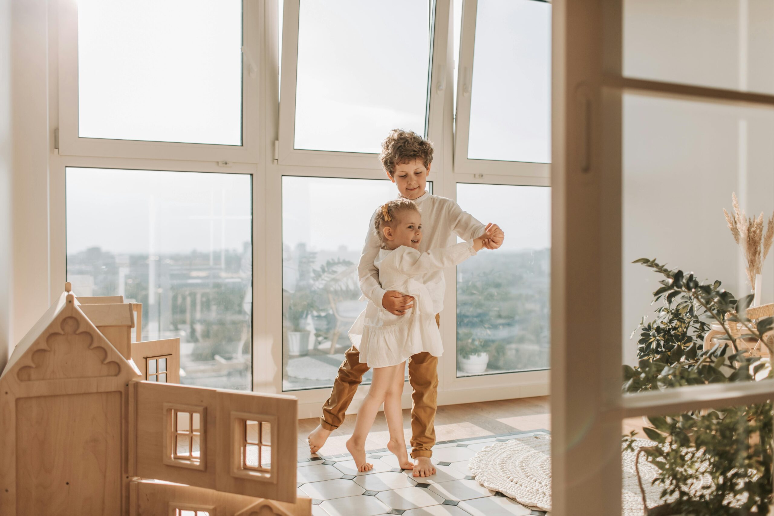 Na slici su dvoje djece koja stoje blizu prozora u svijetloj i sunčanoj sobi. Djeca su okrenuta prema prozoru i plešu.