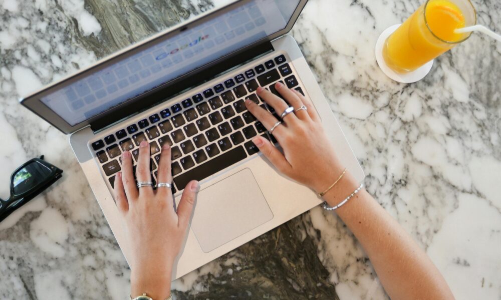 Ova slika prikazuje osobu koja tipka na laptopu. Laptop je postavljen na mramorni stol, a pored njega se nalazi čaša soka od naranče sa slamkom. Osoba nosi sat i nekoliko prstena.