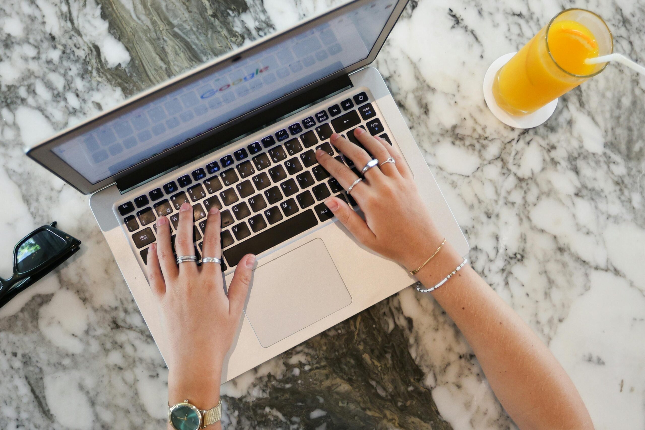 Ova slika prikazuje osobu koja tipka na laptopu. Laptop je postavljen na mramorni stol, a pored njega se nalazi čaša soka od naranče sa slamkom. Osoba nosi sat i nekoliko prstena.