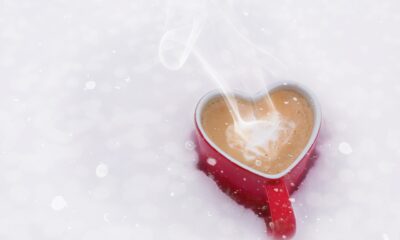 Ova slika prikazuje crvenu šalicu kave u obliku srca koja je postavljena na snijeg. Iz šalice se diže para, stvarajući topao i ugodan kontrast hladnom, snježnom okruženju.