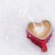 Ova slika prikazuje crvenu šalicu kave u obliku srca koja je postavljena na snijeg. Iz šalice se diže para, stvarajući topao i ugodan kontrast hladnom, snježnom okruženju.