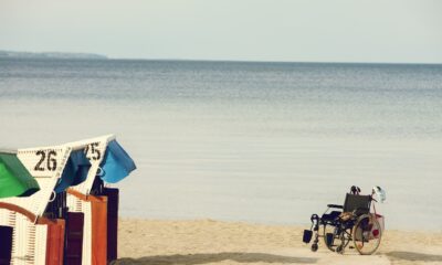Ova slika prikazuje mirnu plažu. Na slici su kabine s brojevima 26 i 25, plave i zelene boje, te invalidska kolica koja su parkirana pored. More je mirno, a horizont je jasno vidljiv.