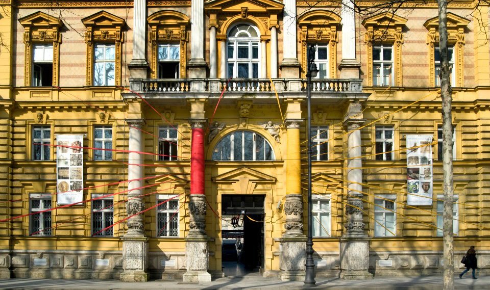 Ova slika prikazuje žutu zgradu s klasičnom arhitekturom. Zgrada ima više prozora, a glavni ulaz je okružen stupovima i ukrašen crvenim trakama. Na zgradi su postavljeni plakati,