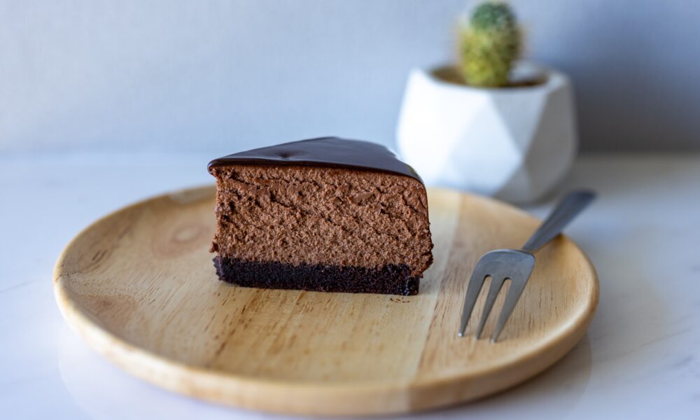 Na slici se nalazi komad čokoladne torte na drvenom tanjuru. Torta ima bogati sloj tamne čokolade na vrhu i izgleda vlažno i gusto. Vilica je postavljena pored komada torte, što ukazuje da je spremna za jelo. Tanjur je okrugao i izrađen je od svijetlog drva.