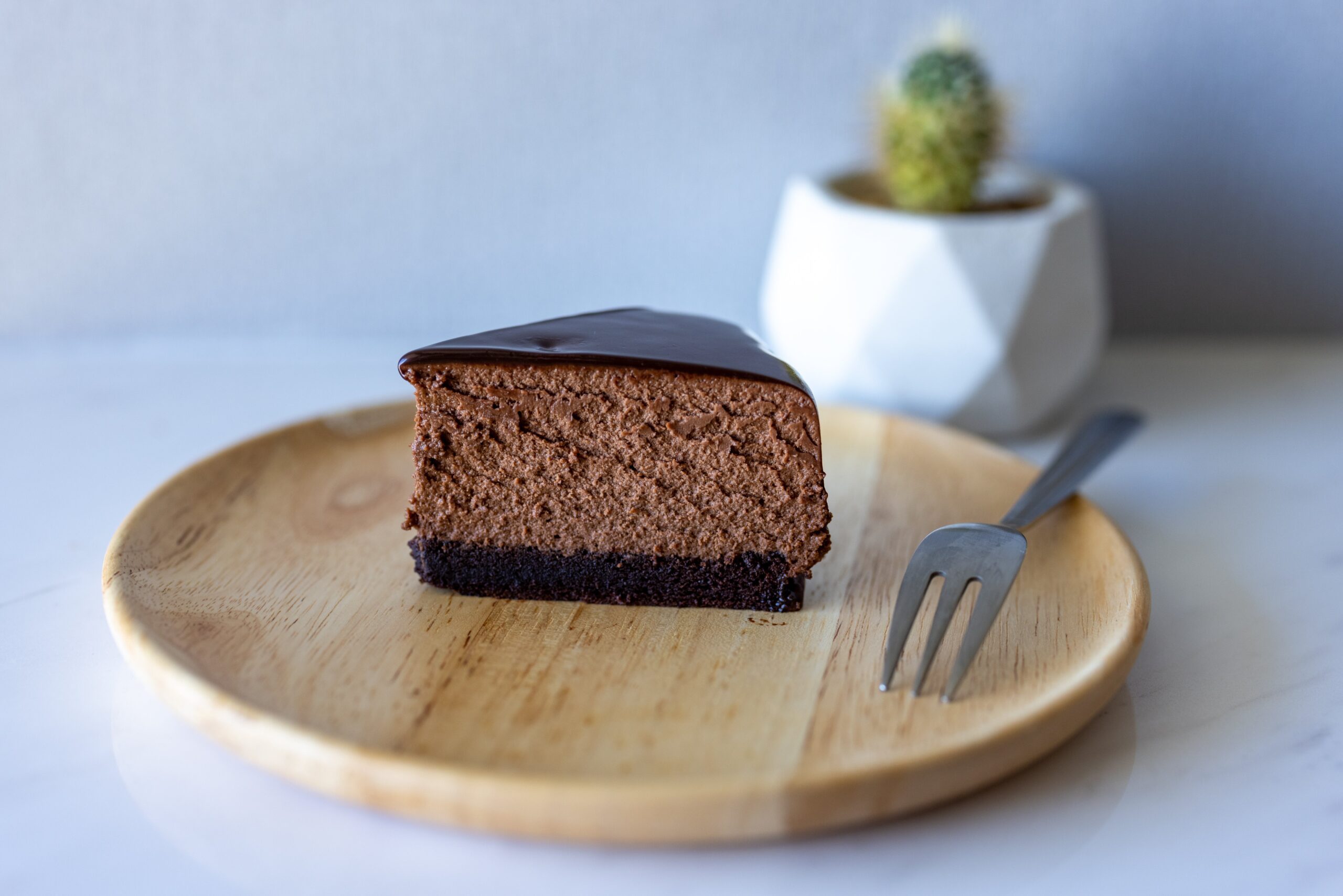Na slici se nalazi komad čokoladne torte na drvenom tanjuru. Torta ima bogati sloj tamne čokolade na vrhu i izgleda vlažno i gusto. Vilica je postavljena pored komada torte, što ukazuje da je spremna za jelo. Tanjur je okrugao i izrađen je od svijetlog drva.