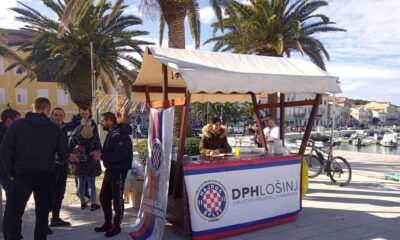 Na štandu stoje dvoje ljudi. Štand ima bijelu tendu, a ispred štanda stoji bijeli plakat na kojem piše "Društvo prijatelja Hajduka Lošinj". Pokraj štanda stoji grupa ljudi.