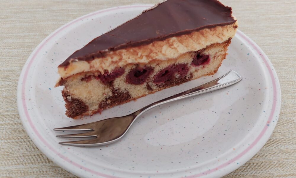 Na slici je komad torte poslužen na ovalnom tanjuru. Torte ima slojeve biskvita i čokolade, a između njih su vidljive crvene bobice, vjerojatno trešnje ili višnje. Također, torta je prelivena glazurom od čokolade na vrhu. Uz tortu na tanjuru se nalazi vilica.