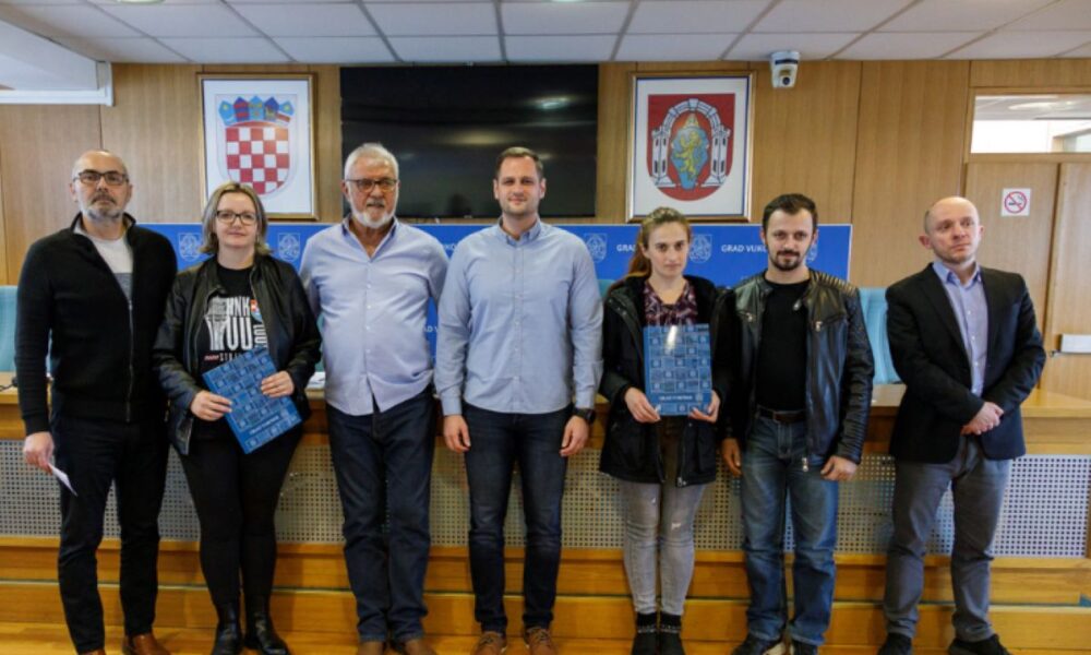 Na fotografiji je skupina ljudi koja stoji ispred pulta s oznakama i grbom. Drže neke dokumente ili certifikate. Iza njih se nalazi pult s raznim oznakama i grbom. Fotografija je snimljena u formalnom okruženju, vjerojatno unutar neke službene ili institucionalne zgrade.