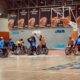 Ova slika prikazuje utakmicu u košarci u invalidskim kolicima. Igrači u plavim i narančastim dresovima natječu se na terenu koji je okružen oglasnim pločama i košem u pozadini. Na zidu iza igrača nalaze se plakati koji prikazuju ime pobjednika raznih košarkaških natjecanja. Turnir se održava u zatvorenom prostoru, a teren je obilježen bijelim linijama. Oglasne ploče i logotipi sponzora su vidljivi na terenu