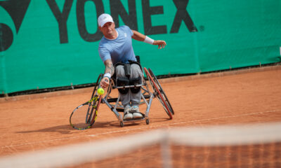 Na slici je osoba u invalidskim kolicima koja igra tenis na crvenoj glinenoj podlozi. Osoba nosi plavu majicu i bijelu kapu. Pokušava udariti žutu tenisku lopticu s reketom. U pozadini je zelena ograda s natpisom “YONEX”.